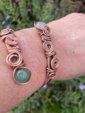Autorska bransoletka miedziana na miłość i relacje prezentująca kwarc różowy oraz awenturyn w autorskim oplocie wire wrapping trzymana w dłoni kobiety na tle kwiatów.